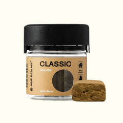 A small 10 Gram jar of Nine Realms Classic Mousse hash with a black lid, shown next to a textured brown bait cube on a light cream background.