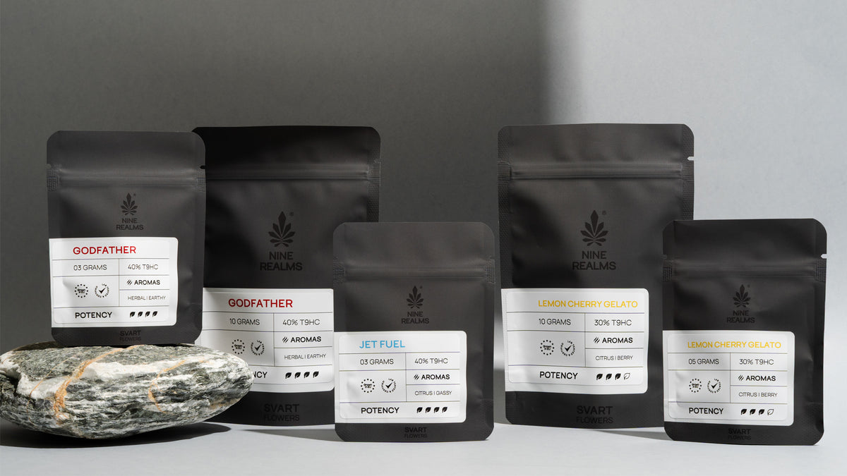 Six matte black stand-up pouches of Nine Realms SVART T9HC Cannabis flowers in various strains and potencies, arranged on a gray surface with a stone slab in the foreground.