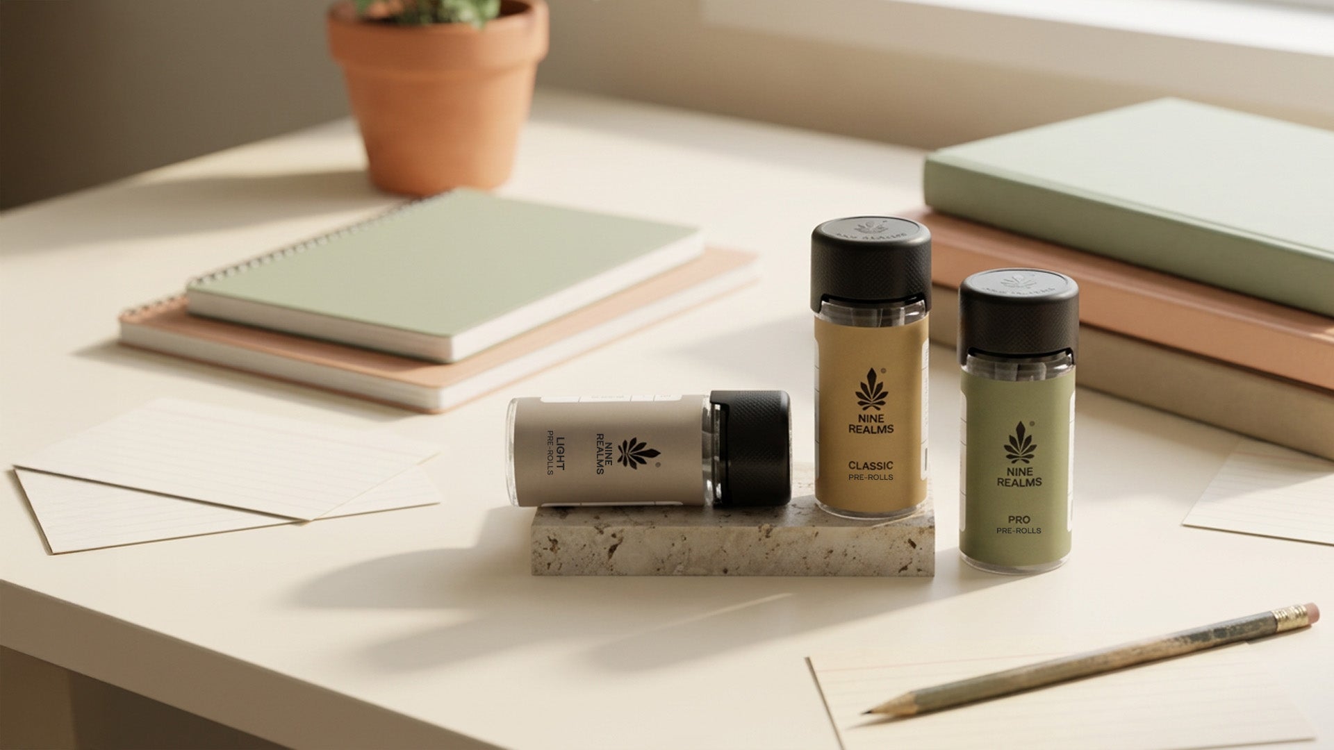 Three Nine Realms THC Joints packages on a light tabletop with notebooks, a pencil, and a plant in the background. The bottles are shown in a styled indoor setting rather than as a plain studio cutout.
