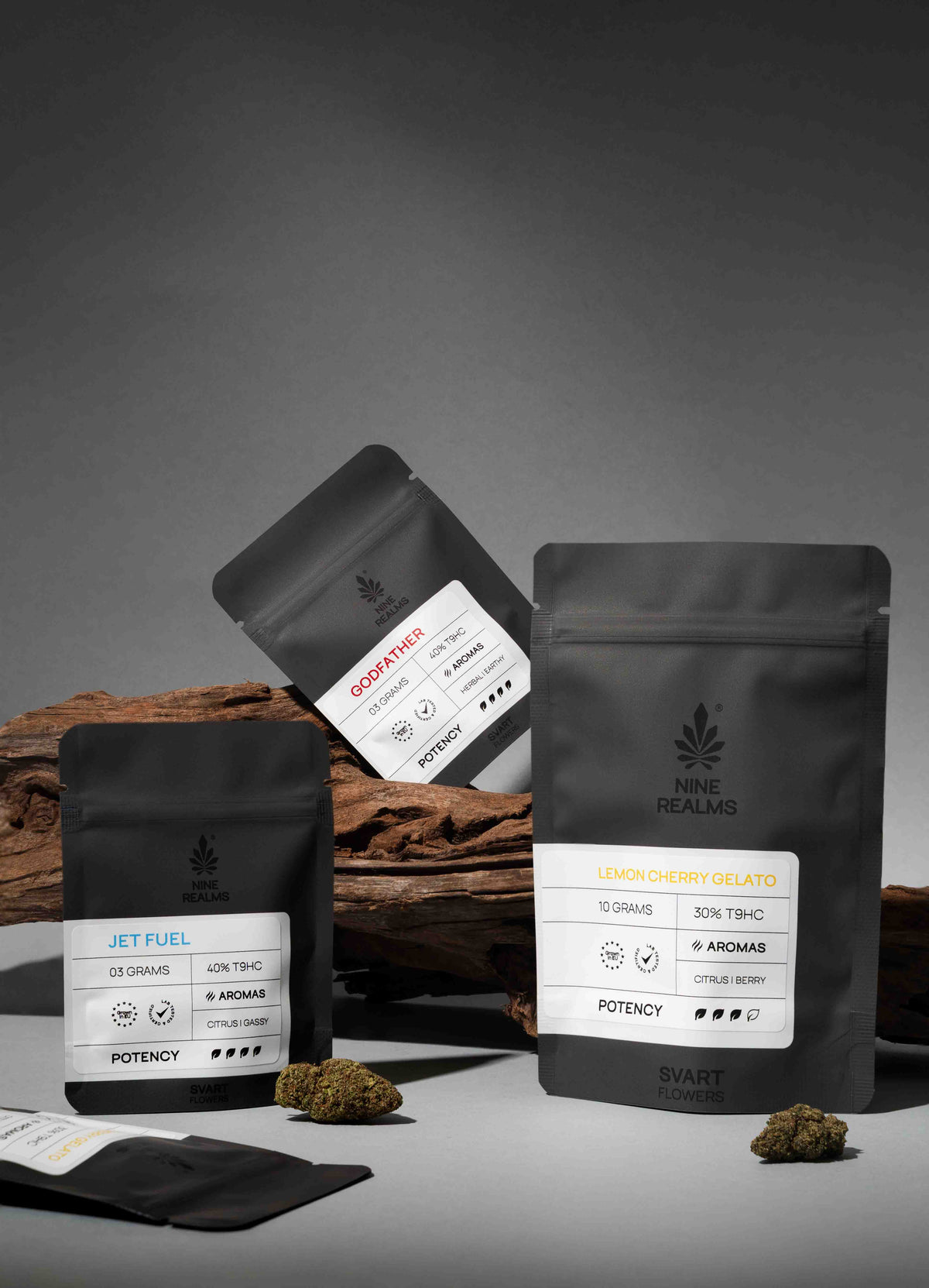 Three matte black stand-up pouches of T9HC cannabis flower from NINE REALMS are arranged on a dark gray studio surface with wood props and small loose buds. The front pouch reads 'LEMON CHERRY GELATO' and the others show 'JET FUEL' and 'GODFATHER'.